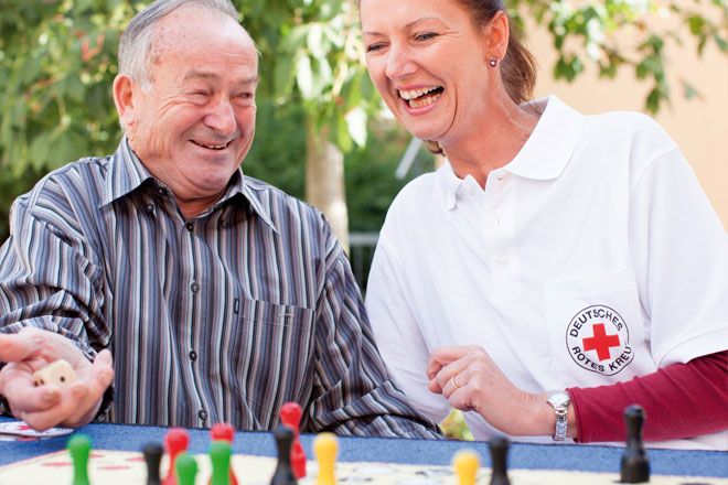 Foto: Eine DRK-Mitarbeiterin spielt 'Mensch ärgere dich nicht' mit einem älteren Herrn. Beide scheinen dabei sichtlich Spaß zu haben und lachen miteinander.