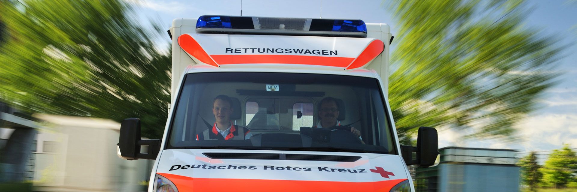 Foto: Zu sehen ist ein DRK-Rettungswagen von vorne in schneller Fahrt. Fahrer und Beifahrer konzentrieren sich auf die Fahrt.