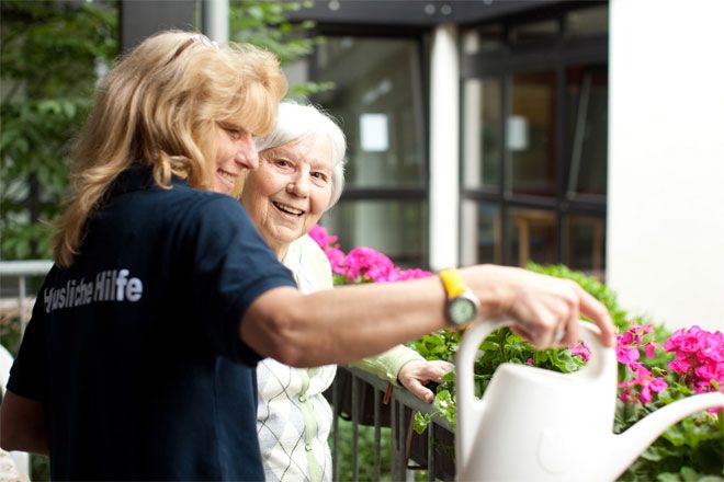 Foto: Eine DRK-Mitarbeiterin gießt die Balkonblumen in der Wohnung einer älteren Dame. Die ältere Dame wirkt glücklich und konzentriert sich mehr auf das gemeinsame Gespräch als auf die Blumen.